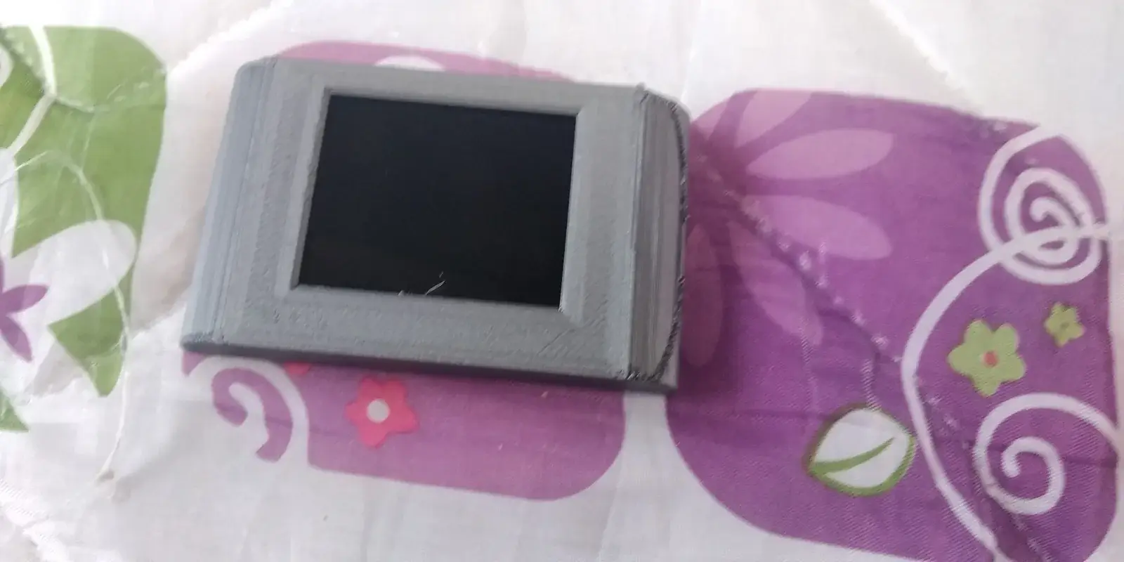 Carcasa gris impresa en 3D con la pantalla TFT montada al frente, apoyada sobre una tela estampada