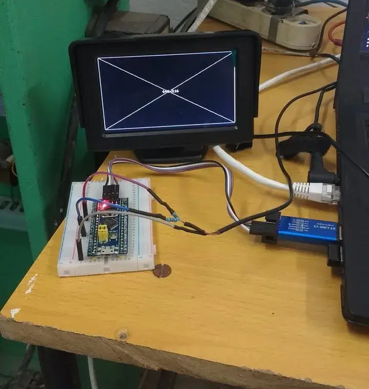 Sobre una mesa de taller, una placa Blue Pill en protoboard conectada por cables a un monitor pequeño que muestra un rectángulo con sus dos diagonales y el texto 448x216 en el centro, con un programador ST-Link azul enchufado a una laptop