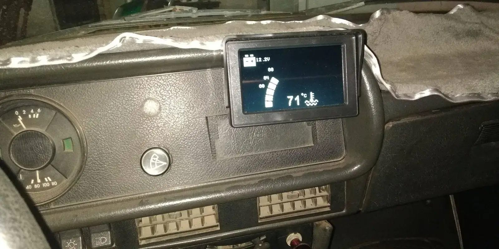 Tablero de un Moskvich con un monitor pequeño montado sobre la consola, encendido con fondo azul oscuro: arriba a la izquierda un icono de batería con 12.2V, al centro una barra de segmentos escalonada con las marcas 80, 84 y 88, y a la derecha el número 71 en grande junto a un icono de termómetro sobre ondas
