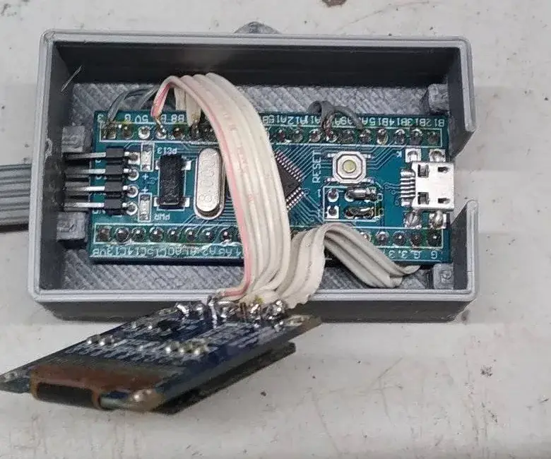 Caja impresa en 3D abierta con una placa STM32 Blue Pill dentro y una pantalla OLED soldada a ella con cables, apoyada fuera de la caja