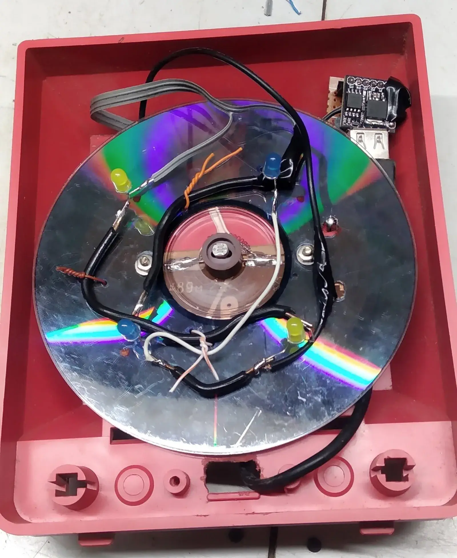 Caja de plástico rojo abierta con un CD viejo dentro como base: sobre el disco, cuatro LEDs sueltos —dos amarillos y dos azules— soldados al aire con cables negros, una pieza redonda transparente al centro con un LDR encima y, arriba a la derecha, una placa negra pequeña con un conector USB hembra
