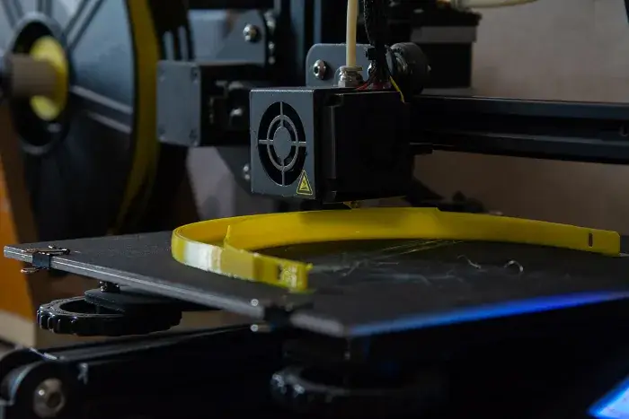Primer plano del extrusor de una impresora 3D depositando filamento amarillo sobre la cama: la diadema de la visera está a medio imprimir, apoyada de canto